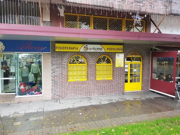 Fachada de la clínica en la Praza Eugenio Fadrique: bajo de ladrillo con ventanas y puerta amarillas y rótulo Fisioterapia Santomé Podología (foto del centro)